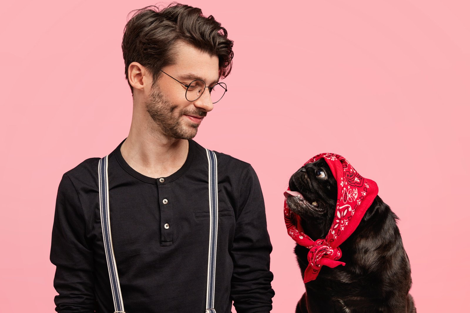 Affectionate male dog owner looks with pleased expression and love at his funny pet with bandana on head, feels responsibility, play together at home, isolated over pink background. Pedigree puppy